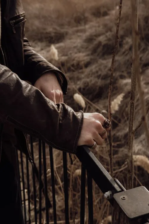 A man wearing rings and a leather jacket beside dry outdoor grasses